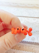 Load image into Gallery viewer, A pair of orange fish stud earrings held between finger and thumb