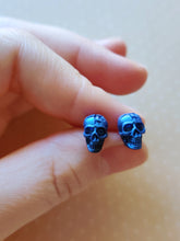 Load image into Gallery viewer, A close of picture of metallic sapphire colored skull earrings held between finger and thumb.