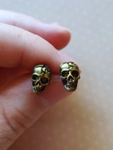 Load image into Gallery viewer, Faux aged gold skull earrings attached to plastic posts, held between finger and thumb.