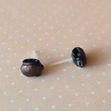 Load image into Gallery viewer, Two miniature flat backed cauldron earrings in black, brushed with a bronze colored metallic pigment. Earrings are displayed on a pale orange background with white polka dots.