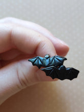 Load image into Gallery viewer, One pair of flat backed bat earrings with relief sculpted detail. Stud earrings are displayed between finger and thumb.