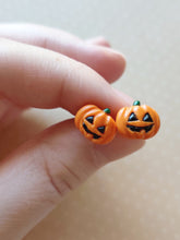 Load image into Gallery viewer, A pair of flat backed jack o' lantern earrings with relief sculpted detail and painted on face and stem. The earrings are held between finger and thumb.