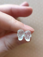 Load image into Gallery viewer, One pair of clear skull stud earrings held between finger and thumb.