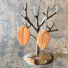 Load image into Gallery viewer, A pair of orange beech leaf shaped polymer clay earrings are displayed hanging from a small, polished silver tree shaped display.
