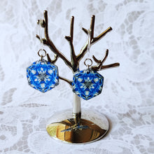 Load image into Gallery viewer, A pair of blue, white, and silver kaleidoscope hexagon earrings hangs from a small polished silver tree stand.