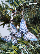 Load image into Gallery viewer, Two white, silver, and blue butterfly earrings with stainless steal jump rings and plastic ear hooks. The earrings are shown hanging from the branch of an artificial Christmas tree.