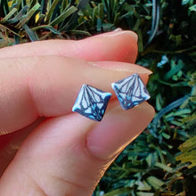 Load image into Gallery viewer, A symmetrical square abstract design of polymer clay earring studs in white, blue and metallic silver held between finger and thumb in front of an artificial Christmas tree.