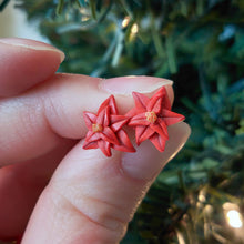 Load image into Gallery viewer, A pair of red Poinsettia earrings held between finger and thumb in front of an artificial Christmas tree.