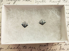 Load image into Gallery viewer, A symmetrical square abstract design of polymer clay earring studs in white, blue and metallic silver held between finger and thumb inside a white paper jewelry box.