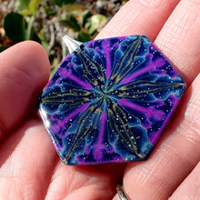 Load image into Gallery viewer, A large hexagonal pendant held in hand in the sunlight. The pendent is dark blue with purple, gold, and light blue colors radiating out from the center. The light reflects off of tiny gold and blue sparkles and small silver glitter. The whole pendant has a shiny gloss finish.