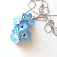 Load image into Gallery viewer, A medium pendant attached to a silver colored chain and bail. The pendant is a cluster of five Forget Me Not flowers. The flowers are pale blue and fade to white toward the center. Each petal ends in yellow at the middle and the center is black. The pendant is displayed on a white ceramic background.