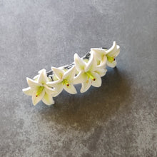 Load image into Gallery viewer, A French style hair barrette with four white lilies made of polymer clay lined up across the top. The barrette has a silver finish and four bezels which each of the lilies are set into. The hair clip is displays on a mottled dark grey surface.