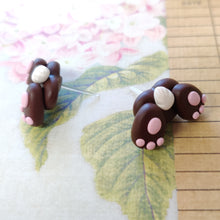 Load image into Gallery viewer, Two small bunny butt shaped earrings resting on a flowered background. They are grey with pink food pads, three toes, and a white tail.