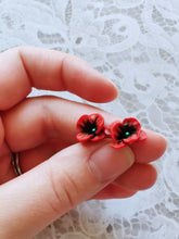 Load image into Gallery viewer, Red and black flower shaped earrings with green metallic centers held between finger and thumb
