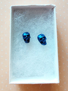 A close of picture of metallic  sapphire colored skull earring in a white paper jewelry box.