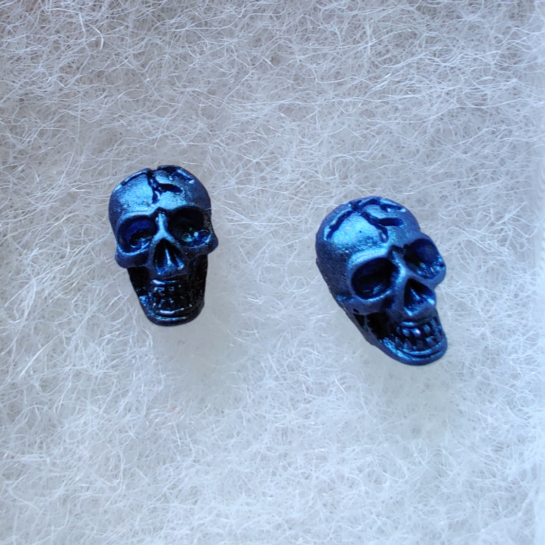 A close of picture of metallic  sapphire colored skull earrings attached to plastic posts. 