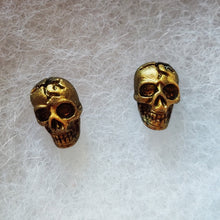 Load image into Gallery viewer, A close of picture of faux aged gold skull earrings attached to plastic posts. 
