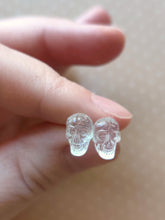 Load image into Gallery viewer, One pair of clear skull stud earrings held between finger and thumb.
