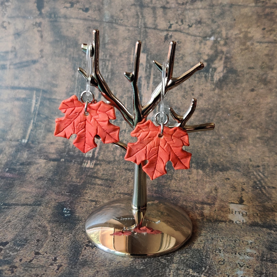 A pair of red maple leaf shaped polymer clay earrings are displayed hanging from a small, polished silver tree shaped display.