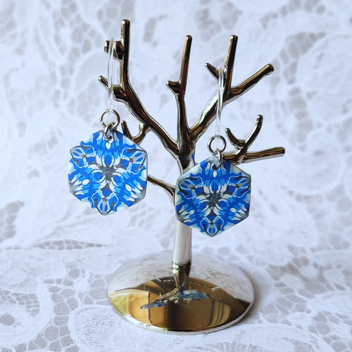 A pair of blue, white, and silver kaleidoscope hexagon earrings hangs from a small polished silver tree stand. 