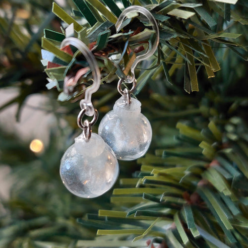 Two miniature Christmas ornament baubles in a clear color with silvery white swirls inside. The earrings are shown hanging from the branch of an artificial Christmas tree. 