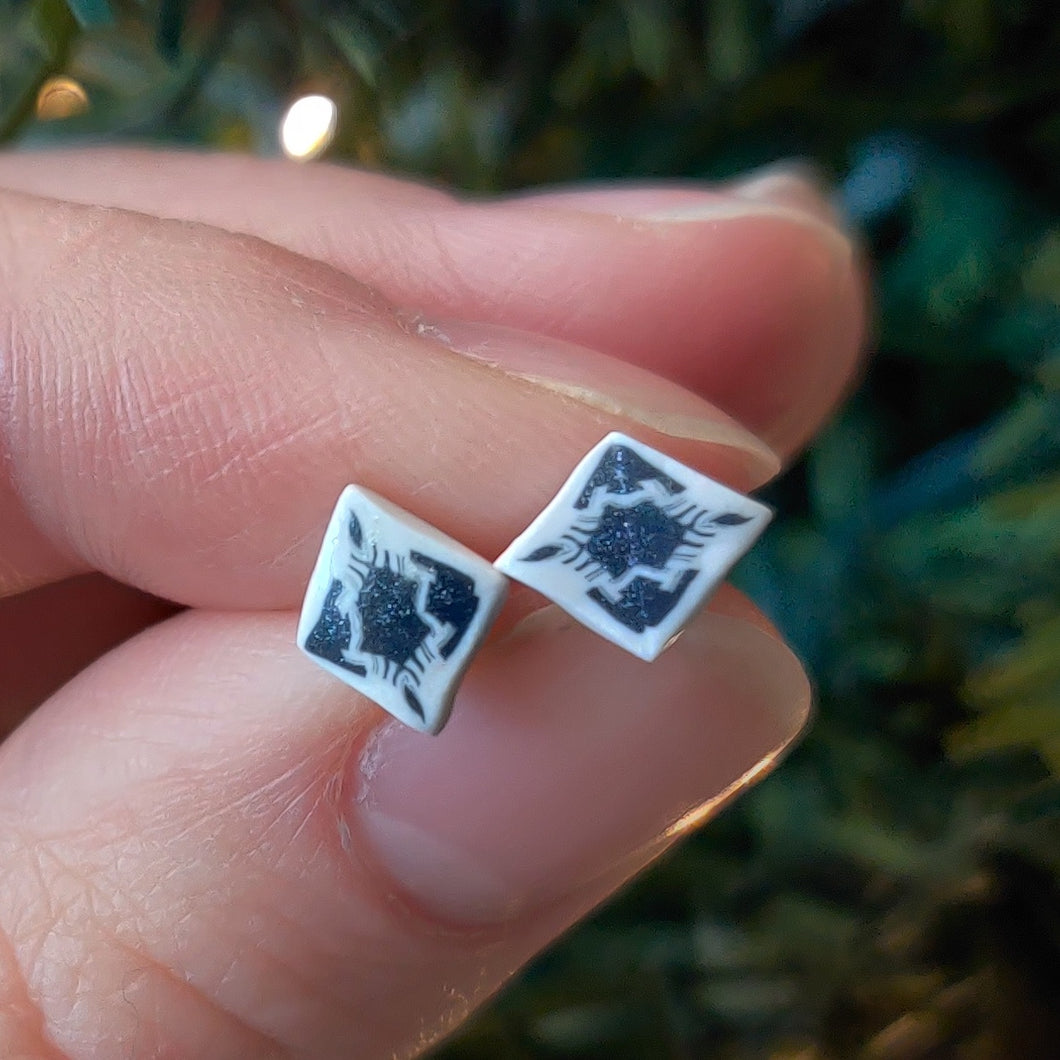 Small, diamond shaped stud earrings  with an abstract white and silver design held between finger and thumb. 