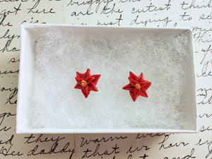 A pair of red poinsettia earrings with red and green centers and darker red veins inside a white paper jewelry box. 