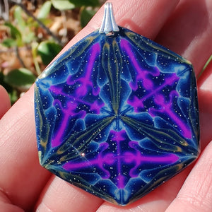 A large hexagonal pendant with a silver colored bail resting in a hand in the sunlight. The pendant is navy blue with veins of gold around the edges and radiating from the center in thirds. There are purple veins in the relative shape of a triangle throughout the pendant. 