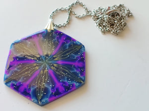 A large hexagonal pendant  connected to a silver colored chain and bail. The pendent is dark blue with purple, gold, and light blue colors radiating out from the center. It is displayed laying on a white plain ceramic tile.