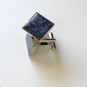 A pair of silver colored cufflinks with a bezel on top. The bezel is filled with polymer clay with a shiny gloss finish. The clay is dark blue with veins of gold, purple, and glow in the dark clay radiating from the center. 