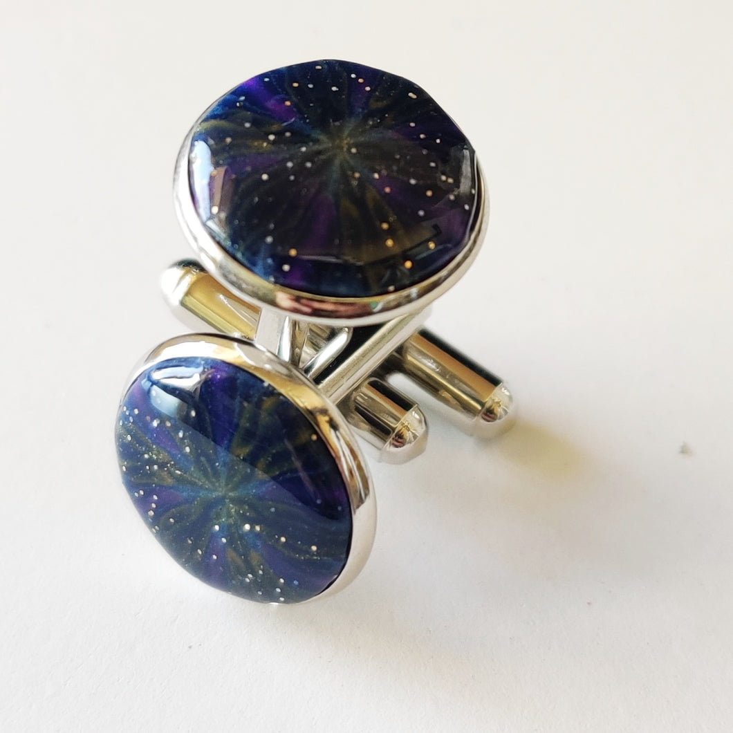 A pair of silver colored cufflinks with a bezel on top. The bezel is filled with polymer clay with a shiny gloss finish. The clay is dark blue with veins of gold, purple, and glow in the dark clay radiating from the center. 