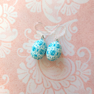 Patterned Easter egg shaped dangle earrings in a blue and white one a pink paper background.